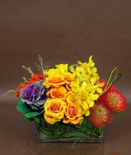 A fascinating arrangement bursting with lush blooms of roses, kale, orchids, and orange pincushion protea. Arranged in a rectangular vase, the array of autumn colors is a visual delight.